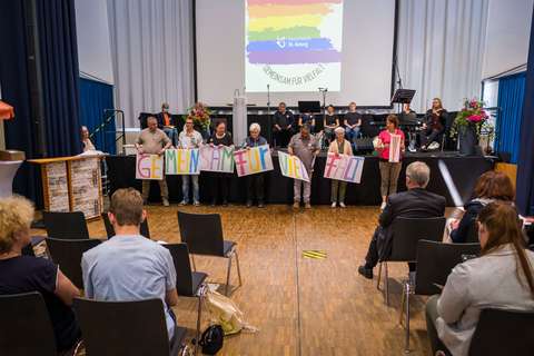 Mehrere Personen stehen in einer Reihe und halten bunte Schilder mit dem Schriftzug „GEMEINSAM FÜR VIELFALT“, während im Hintergrund weitere Personen auf einer Bühne sitzen und Musikinstrumente zu sehen sind.