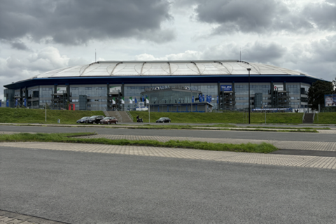 Ein großes Stadiongebäude mit blauer Fassade und weißem Dach, davor mehrere Autos und eine Person auf einem Motorrad.