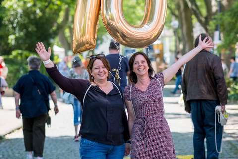 Zwei Personen stehen auf einer Straße und halten große, goldene Luftballons in Form der Zahl 70.