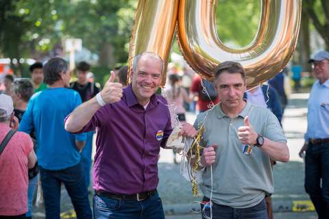 Zwei Personen stehen nebeneinander im Freien, halten goldene Luftballons und zeigen den Daumen nach oben.