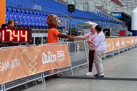 Eine Person im Tierkostüm und zwei Personen geben sich an einer Absperrung in einem Stadion die Hand.