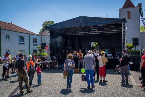 Mehrere Personen stehen auf einem gepflasterten Platz vor einer Bühne im Freien.