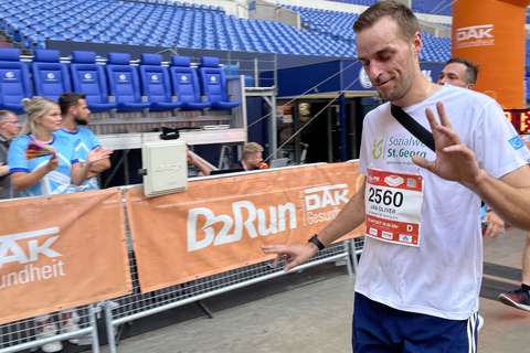 Eine Person läuft in Sportkleidung mit Startnummer an einer Ziellinie in einem Stadion vorbei, im Hintergrund stehen weitere Personen und leere Sitzreihen.