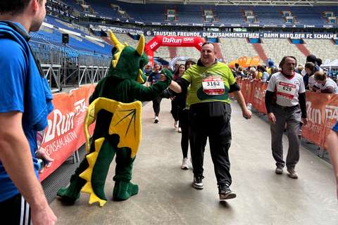 Eine Person im Drachenkostüm gibt einer Person mit Startnummer 3162 ein High-Five, während mehrere Personen an einem Laufwettbewerb in einem Stadion teilnehmen.