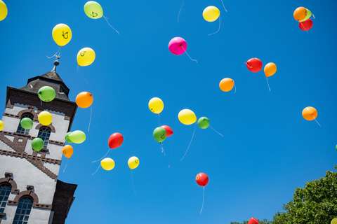 Mehrere bunte Luftballons steigen vor einem Kirchturm und blauem Himmel auf.
