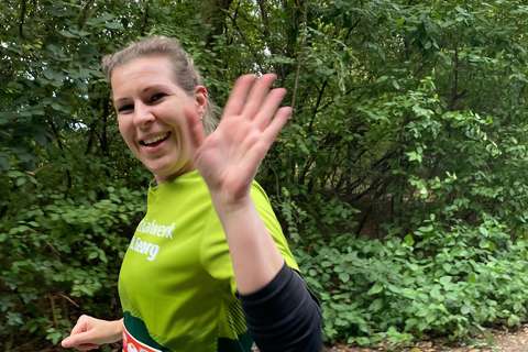 Eine Person mit grünem Laufshirt läuft im Freien und winkt in die Kamera, im Hintergrund sind dichte grüne Büsche.