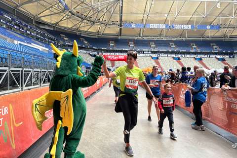 Eine Person im grünen Drachenkostüm gibt einer laufenden Person ein High-Five, weitere Personen laufen und stehen in einem Stadion.