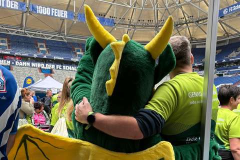 Eine Person im grünen Drachenkostüm mit gelben Hörnern umarmt eine Person in grünem T-Shirt in einem Stadion.