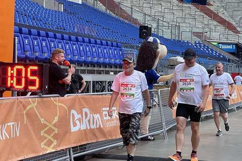 Drei Personen mit Startnummern gehen in einem Stadion an einer Ziellinie vorbei, daneben steht eine Person im Maskottchenkostüm.
