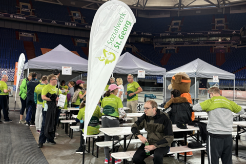 Mehrere Personen in grünen T-Shirts stehen und sitzen an Bierzeltgarnituren unter weißen Pavillons in einer großen Halle, daneben ein Aufsteller mit der Aufschrift „Sozialwerk St. Georg“.