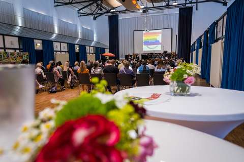Mehrere Personen sitzen in einem Saal und blicken auf eine Bühne mit Leinwand, im Vordergrund steht ein runder Tisch mit einem Blumenstrauß.