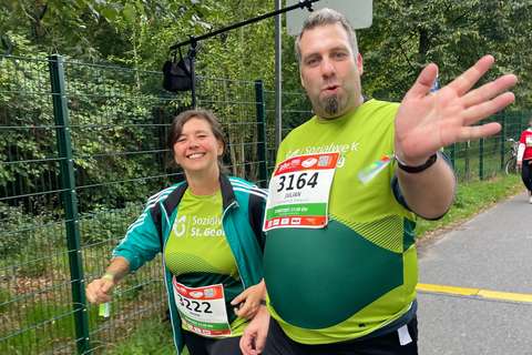 Zwei Personen mit Startnummern und grünen Laufshirts gehen nebeneinander auf einem Weg im Freien, eine Person winkt in die Kamera.