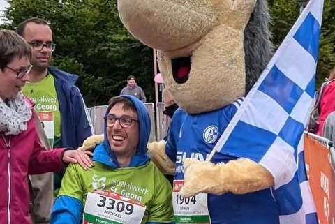 Eine Person im Rollstuhl hält eine Startnummer und wird von einer weiteren Person sowie einem Maskottchen mit blau-weißer Fahne begleitet.