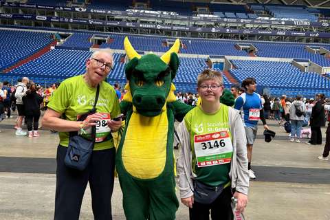 Zwei Personen in grünen T-Shirts und Startnummern stehen neben einer Person im Drachenkostüm in einem Stadion.
