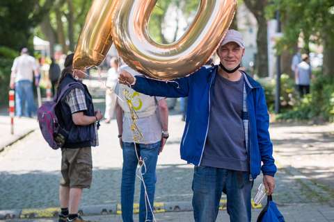 Eine Person hält einen großen, goldenen Luftballon in Form der Zahl 70 und trägt eine Jacke sowie eine Kappe.