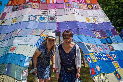 Zwei Personen stehen unter einem großen, bunten Zelt aus gehäkelten oder gestrickten Quadraten auf einer Wiese.