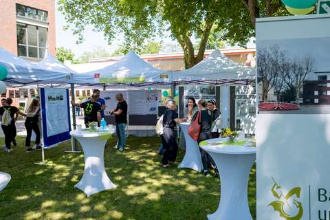 Mehrere Personen stehen und unterhalten sich an Stehtischen vor weißen Pavillons mit Informationspostern im Freien.