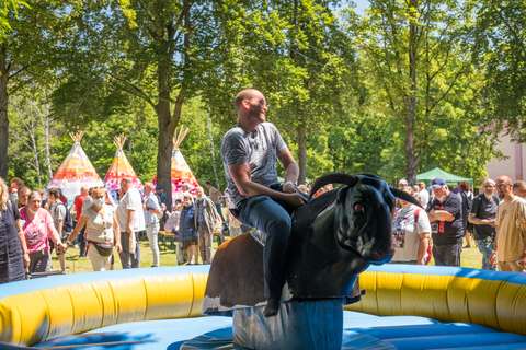 Eine Person reitet auf einem mechanischen Bullen, umgeben von einer Menschenmenge im Freien mit Bäumen und bunten Tipis im Hintergrund.
