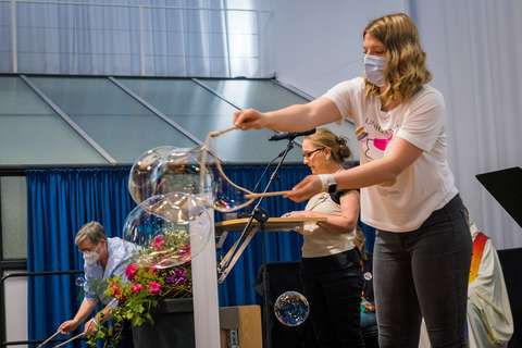 Drei Personen mit Masken, eine Person macht Seifenblasen, eine steht an einem Rednerpult, eine beugt sich über einen Blumenschmuck.