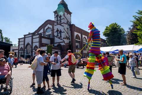 Mehrere Personen stehen und gehen auf einem Platz vor einem großen Gebäude, eine Person auf Stelzen trägt ein buntes Kostüm.
