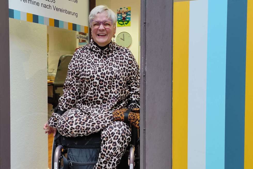 Eine Person im Rollstuhl trägt einen Anzug mit Leopardenmuster und sitzt in einem Eingang neben einer Glastür mit bunten Streifen.