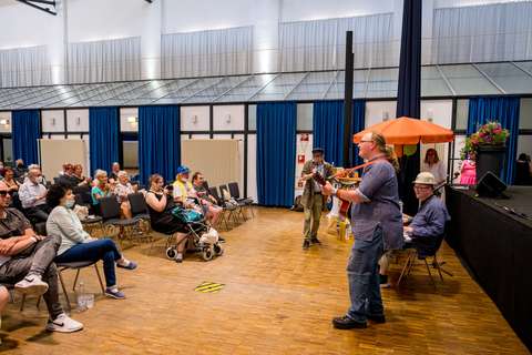 Mehrere Personen sitzen auf Stühlen in einem Saal, während eine Person Gitarre spielt und eine weitere Person am Keyboard sitzt; im Hintergrund steht ein orangefarbener Sonnenschirm und ein großer Blumenstrauß.