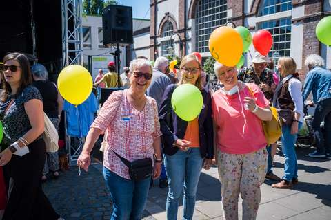 Drei Personen stehen nebeneinander und halten bunte Luftballons, umgeben von weiteren Personen im Freien.