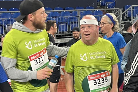 Zwei Personen in Sportkleidung mit Startnummern stehen nebeneinander in einer Halle, umgeben von weiteren laufenden Personen.