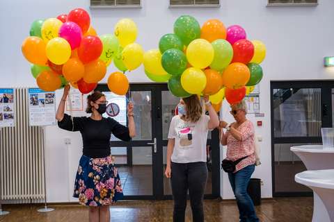Drei Personen stehen in einem Raum und halten viele bunte Luftballons.