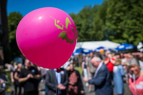 Ein pinker Luftballon mit einem grünen Drachenmotiv schwebt im Vordergrund, im Hintergrund stehen mehrere Personen unscharf auf einer Veranstaltung im Freien.