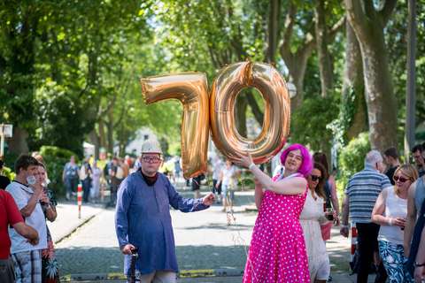 Zwei Personen halten goldene Luftballons in Form der Zahl 70, eine Person trägt ein pinkes Kleid mit weißen Punkten und eine pinke Perücke, im Hintergrund stehen mehrere weitere Personen auf einer von Bäumen gesäumten Straße.