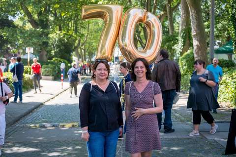 Zwei Personen stehen auf einer Straße vor goldenen Luftballons in Form der Zahl 70, im Hintergrund sind weitere Personen und Bäume.
