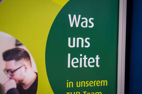 Ein Plakat mit dem Text „Was uns leitet“ und einem teilweise sichtbaren Bild einer Person mit Brille.
