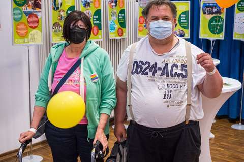 Zwei Personen mit Mund-Nasen-Schutz stehen in einem Raum, eine Person hält einen Rollator und eine gelbe Luftballon, im Hintergrund bunte Infotafeln und weitere Luftballons.