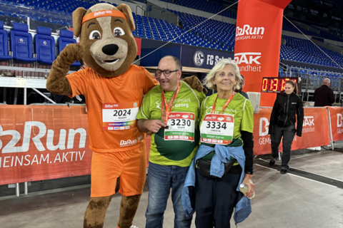 Drei Personen, darunter eine im Hundekostüm, stehen mit Medaillen und Startnummern vor einem roten Zielbogen in einem Stadion.