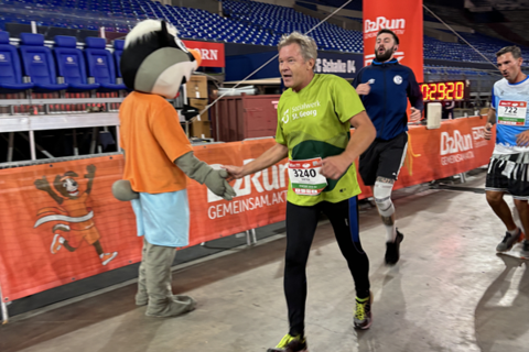 Eine Person im grünen Laufshirt gibt einer als Tier verkleideten Person die Hand, während zwei weitere Personen hinter ihnen laufen und ein roter Zielbogen im Hintergrund steht.