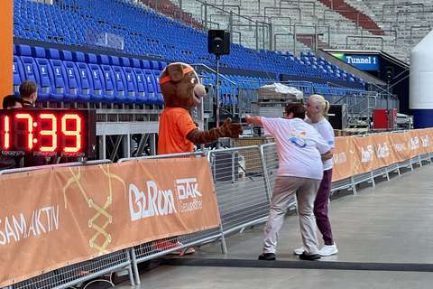 Eine Person im Hundekostüm gibt einer anderen Person die Hand, während eine weitere Person daneben steht; im Hintergrund leere Tribünen in einem Stadion.