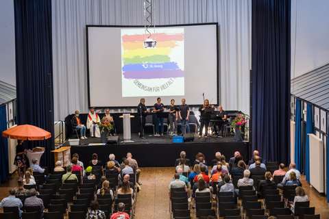 Mehrere Personen sitzen in einem Saal und blicken auf eine Bühne, auf der mehrere Personen stehen und musizieren oder sprechen; im Hintergrund ist eine Leinwand mit einer Regenbogenfahne und dem Text „Gemeinsam für Vielfalt“.