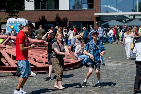 Mehrere Personen stehen und gehen auf einem gepflasterten Platz, im Hintergrund sind weitere Menschen und ein Gebäude zu sehen.