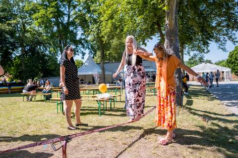 Drei Personen stehen auf einer Wiese, zwei balancieren gemeinsam auf einem Slackline-Band, eine weitere Person steht daneben; im Hintergrund Bänke, Tische und Bäume.