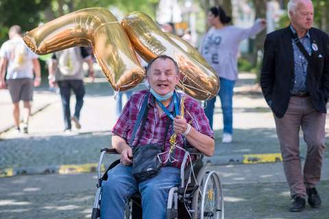 Eine Person im Rollstuhl hält goldene Luftballons in Form der Zahl 70, mehrere Personen gehen im Hintergrund auf einem baumbestandenen Weg.