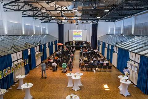 Mehrere Personen sitzen auf Stühlen in einem großen Saal, vorne befindet sich eine Bühne mit einer Leinwand, im Vordergrund stehen runde Stehtische.