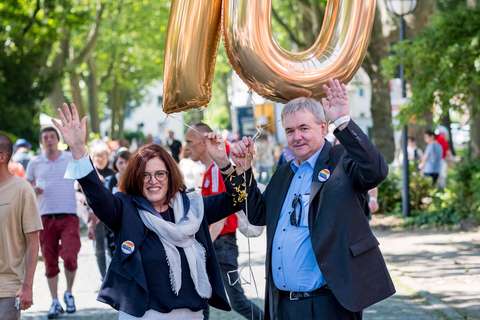Zwei Personen halten goldene Luftballons in Form der Zahl 70 und heben die Arme, im Hintergrund gehen weitere Personen auf einer von Bäumen gesäumten Straße.