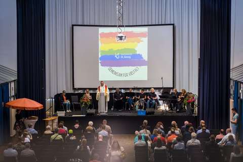 Mehrere Personen sitzen und stehen auf einer Bühne vor einem Publikum, im Hintergrund eine Leinwand mit Regenbogenfarben und der Aufschrift „Sozialwerk St. Georg – Gemeinsam für Vielfalt“.