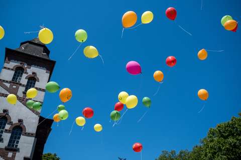 Mehrere bunte Luftballons steigen vor blauem Himmel neben einem Kirchturm auf.