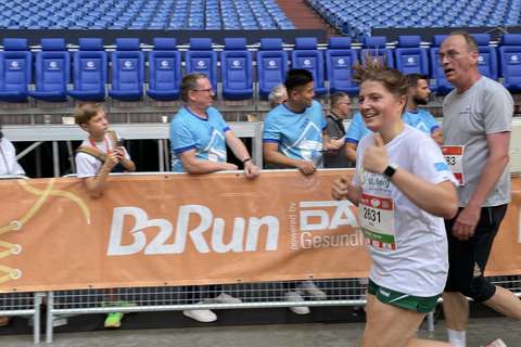 Eine Person läuft in Sportkleidung an einer Absperrung mit „B2Run“-Banner vorbei, im Hintergrund sitzen und stehen mehrere Personen auf Stadionplätzen.