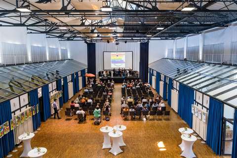 Mehrere Personen sitzen in einem großen Saal auf Stühlen und blicken auf eine Bühne mit Podium und Leinwand, an den Seiten stehen Stehtische und es hängen Bilder an den Wänden.