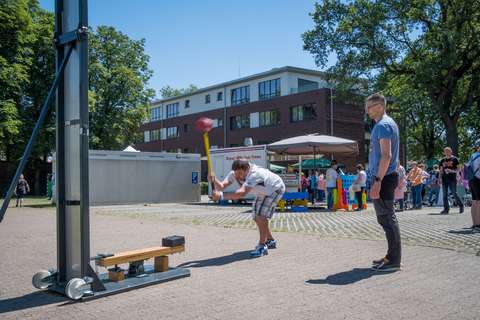 Eine Person schlägt mit einem Hammer auf einen Hau-den-Lukas, während eine weitere Person daneben steht und mehrere Personen im Hintergrund zu sehen sind.