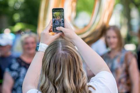 Eine Person mit schulterlangen, blonden Haaren fotografiert mit einem Smartphone eine Gruppe von Personen vor einer großen, goldenen Ballonzahl 70.