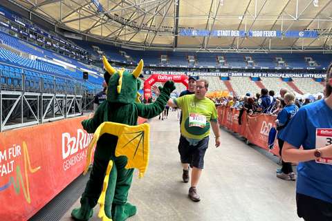 Eine Person im Drachenkostüm gibt einer laufenden Person ein High-Five in einem Stadion, während weitere Personen am Rand stehen.
